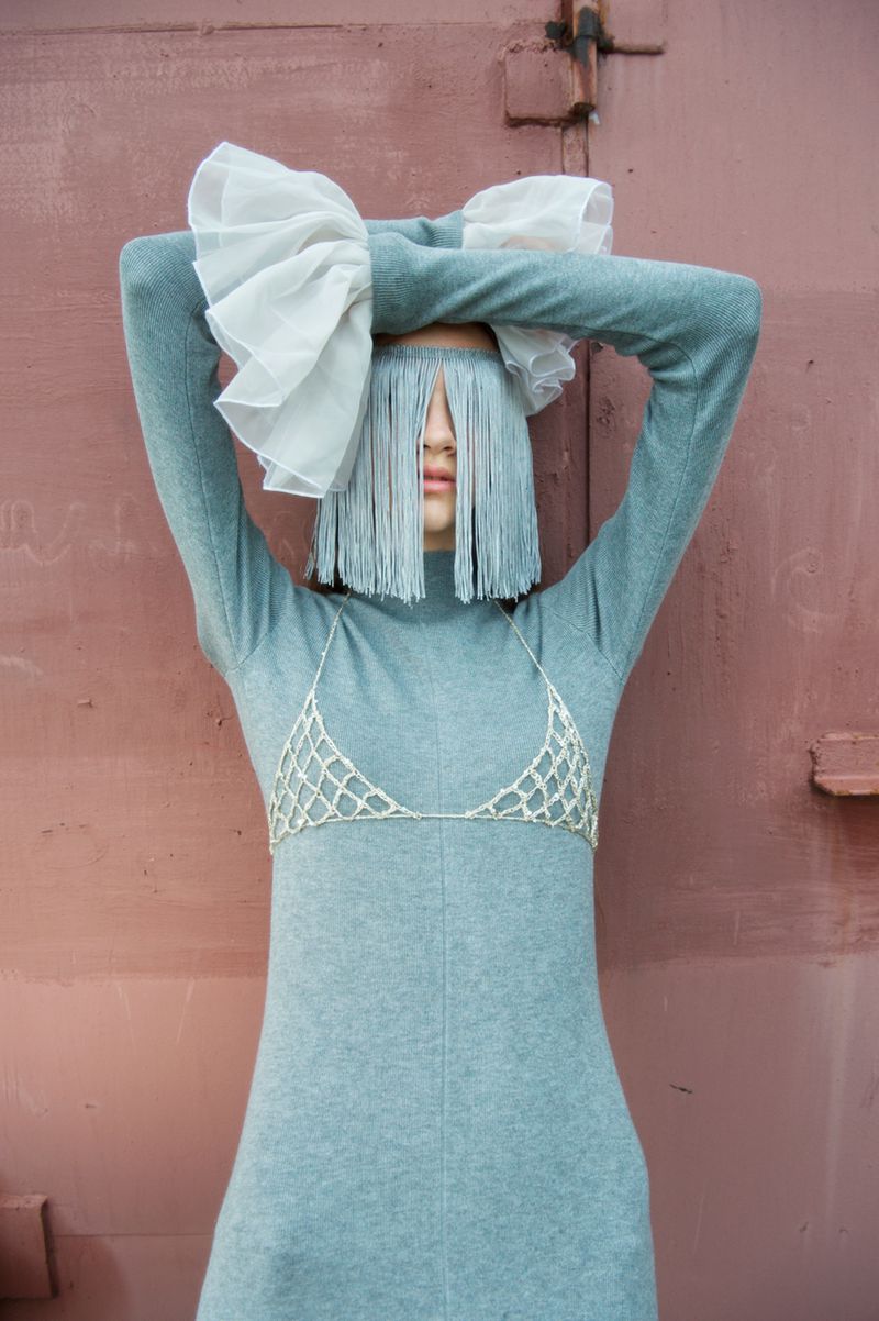 Model wearing a blue sweater dress, beaded bralette, and fringe face covering with white ruffles, standing against a dusty pink wall.