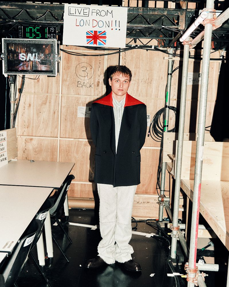 Young man in a black blazer with red shoulder accents and white textured trousers standing backstage by scaffolding and a video monitor.