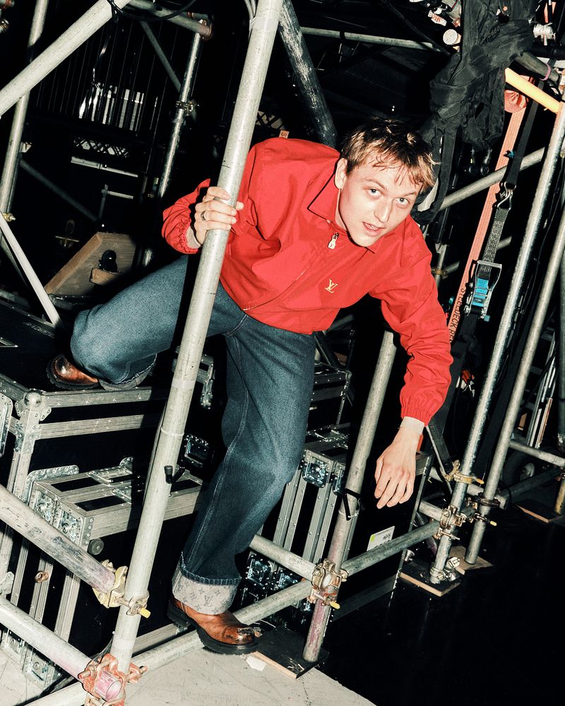 Young man in a vibrant red windbreaker and dark blue jeans climbing through metal scaffolding in a studio environment.