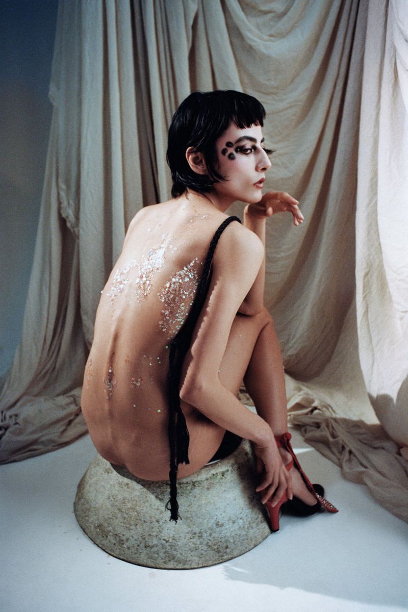 Artistic nude studio shot of an adult woman with face dots sitting on a bowl, wearing red heels with a draped curtain backdrop.