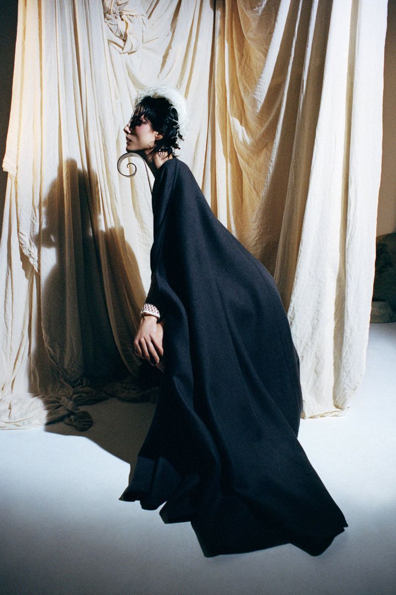 Fashion shot of a woman wearing a large black dress with statement earrings posing in front of draped light fabric in a studio setting.