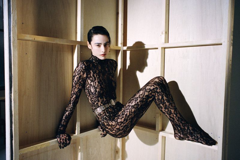 A slender woman in a black lace bodysuit poses artistically against a plywood backdrop in a studio setting, lit with warm lighting.