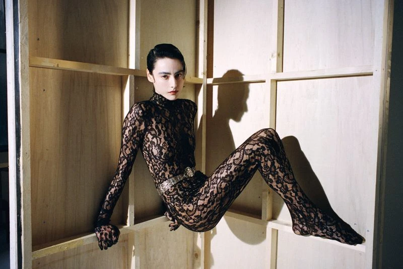 A slender woman in a black lace bodysuit poses artistically against a plywood backdrop in a studio setting, lit with warm lighting.