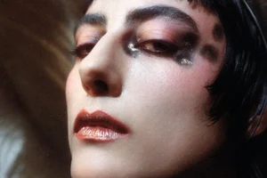 Close-up portrait of a woman with dramatic dark eye makeup and shimmering metallic red lips against a neutral background.