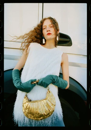 Model Emese Nyiro wearing long green leather gloves and a white fringe top holds a metallic gold crescent handbag.