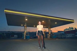 Model Emese Nyiro in a pale yellow polo shirt and black mini skirt standing beneath an illuminated gas station canopy at dusk.