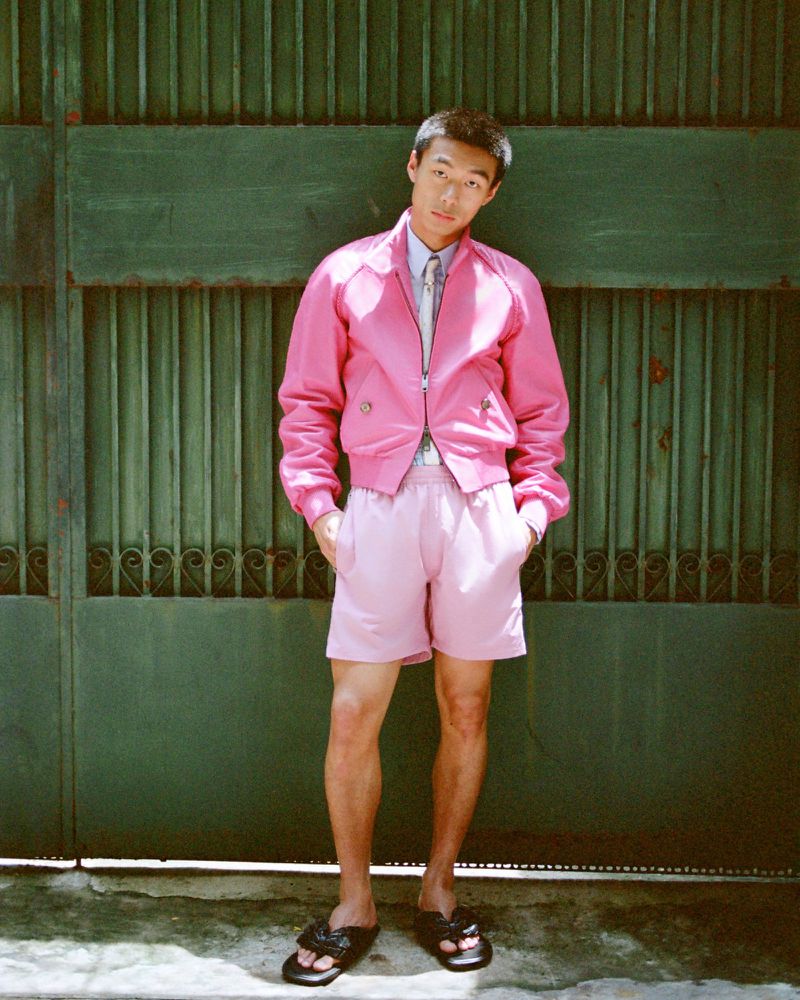 Man wearing a pink jacket and pink shorts posing in front of a green slatted gate for Esquire China editorial