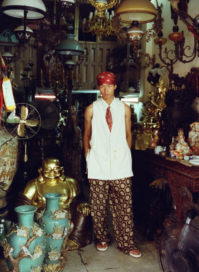 Model Yan Li wearing a sleeveless white vest and patterned trousers stands inside a crowded antique shop surrounded by lamps and statues.