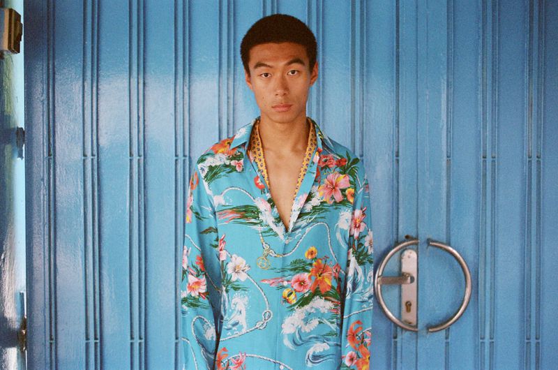Model Yan Li wearing a light blue tropical floral print shirt, standing in front of a bright blue paneled door with a circular handle.