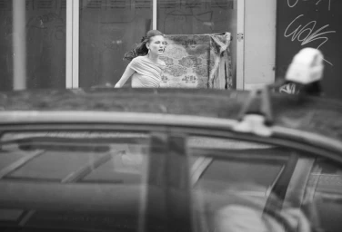 Black and white photo of Nadine Kirilova in a draped top framed through a car foreground on an urban street.