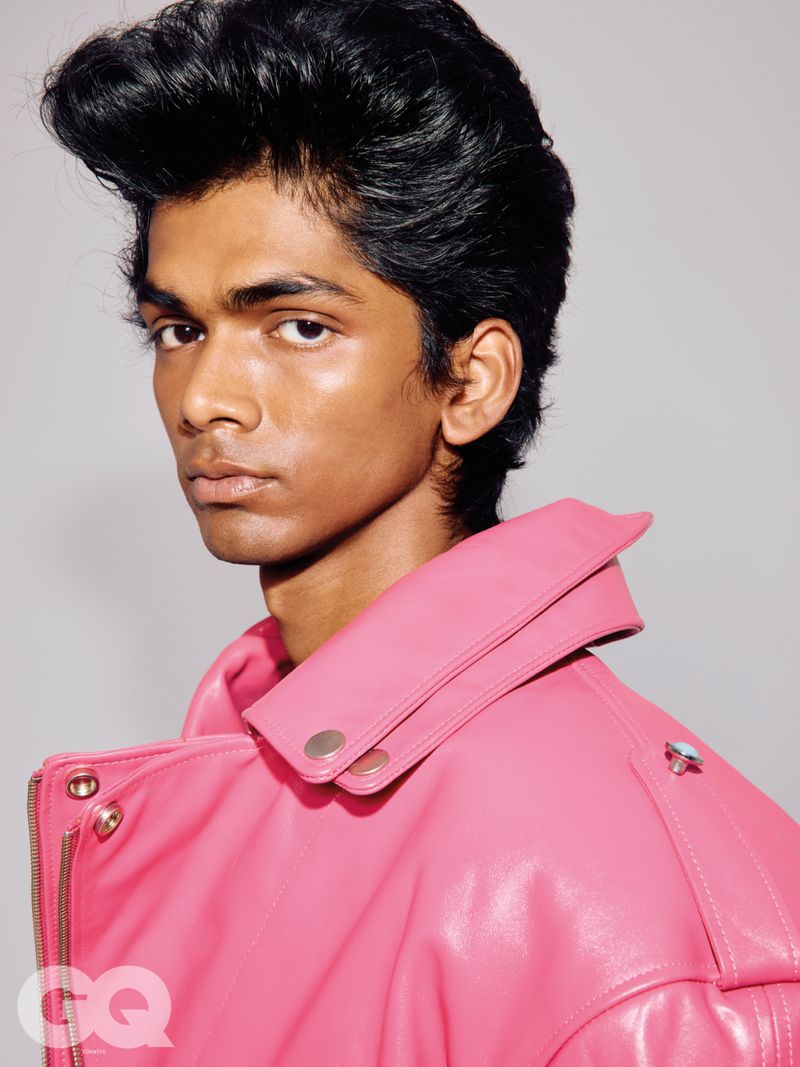 Close-up portrait of a young man with styled black pompadour hair wearing a bright pink leather jacket against a grey background.