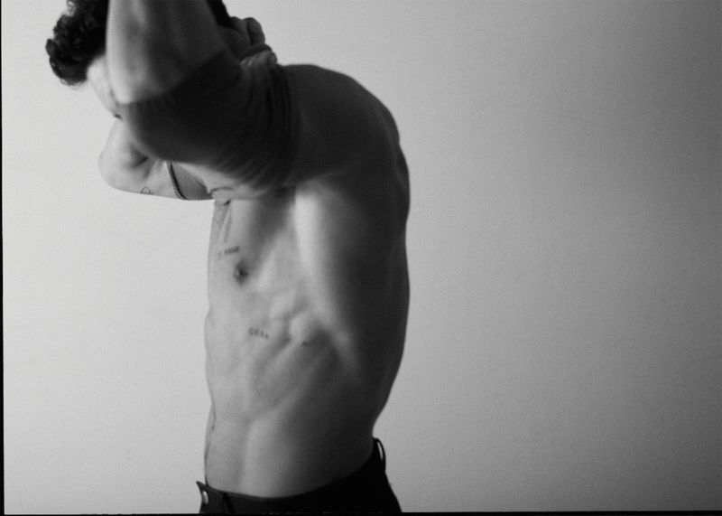 Monochrome, shirtless adult male with defined musculature and tattoos poses in front of a plain light wall.