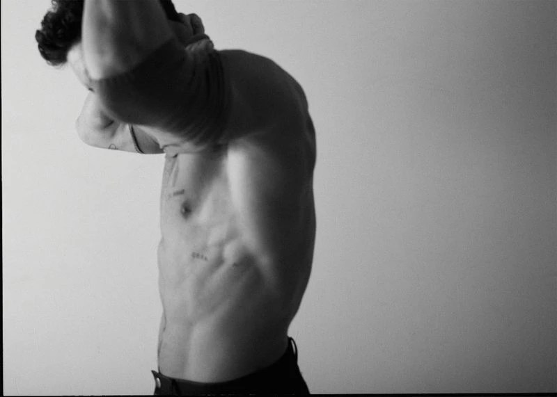 Monochrome, shirtless adult male with defined musculature and tattoos poses in front of a plain light wall.