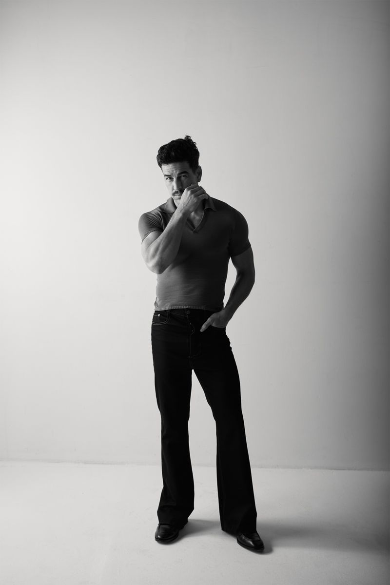 Monochrome studio shot of a man with dark hair and a mustache, wearing a polo shirt and dark jeans, standing against a plain background.