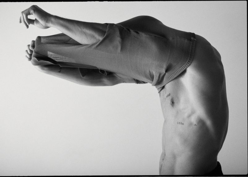 Monochrome studio image of a partially visible adult male figure, bent over with his upper torso visible, wearing jeans pulled over his head.