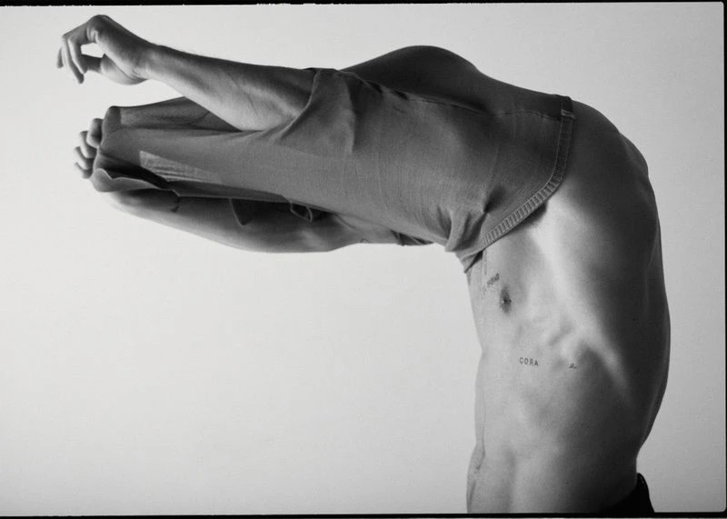 Monochrome studio image of a partially visible adult male figure, bent over with his upper torso visible, wearing jeans pulled over his head.