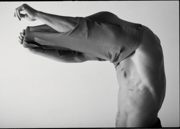 Monochrome studio image of a partially visible adult male figure, bent over with his upper torso visible, wearing jeans pulled over his head.