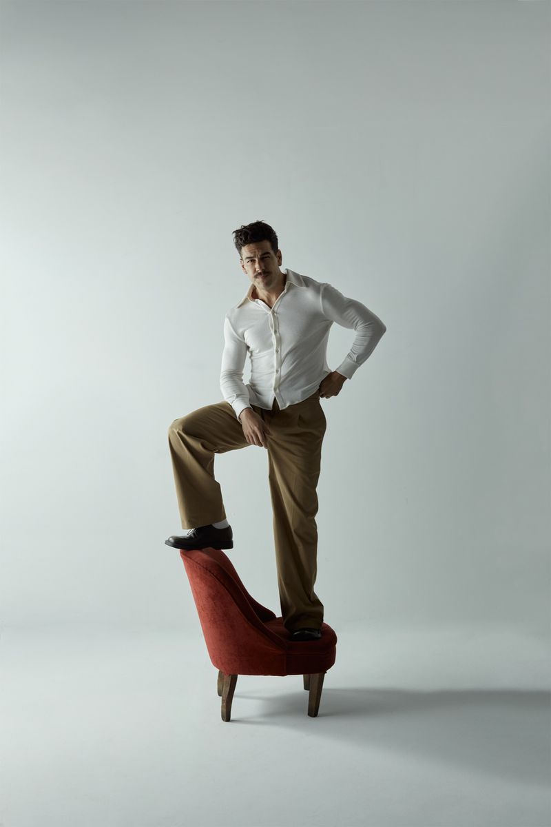 Man in white shirt and tan pants stands on a red chair against a white background in a fashion style portrait, indoor studio shot.