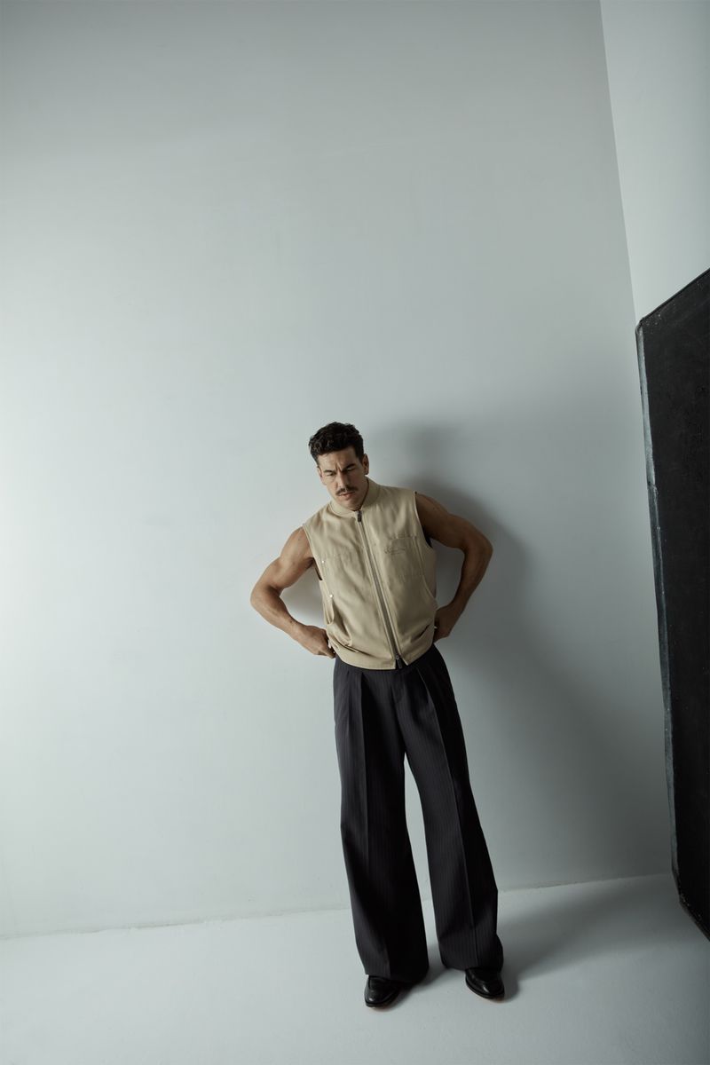 An adult male model poses indoors wearing a sleeveless light brown suede vest, dark trousers, and black shoes against a neutral wall.