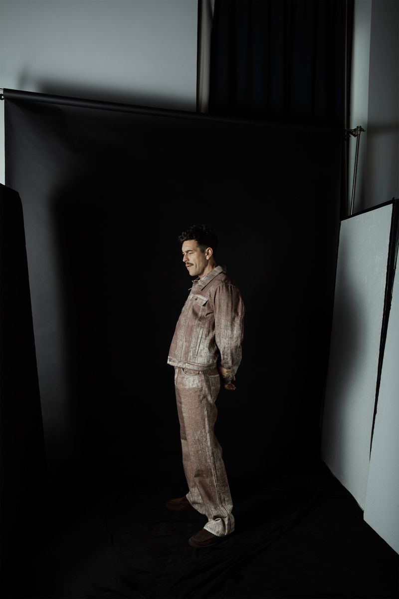 Man poses in a taupe-colored jacket and pants in a photo studio against a black backdrop with studio lighting and equipment.