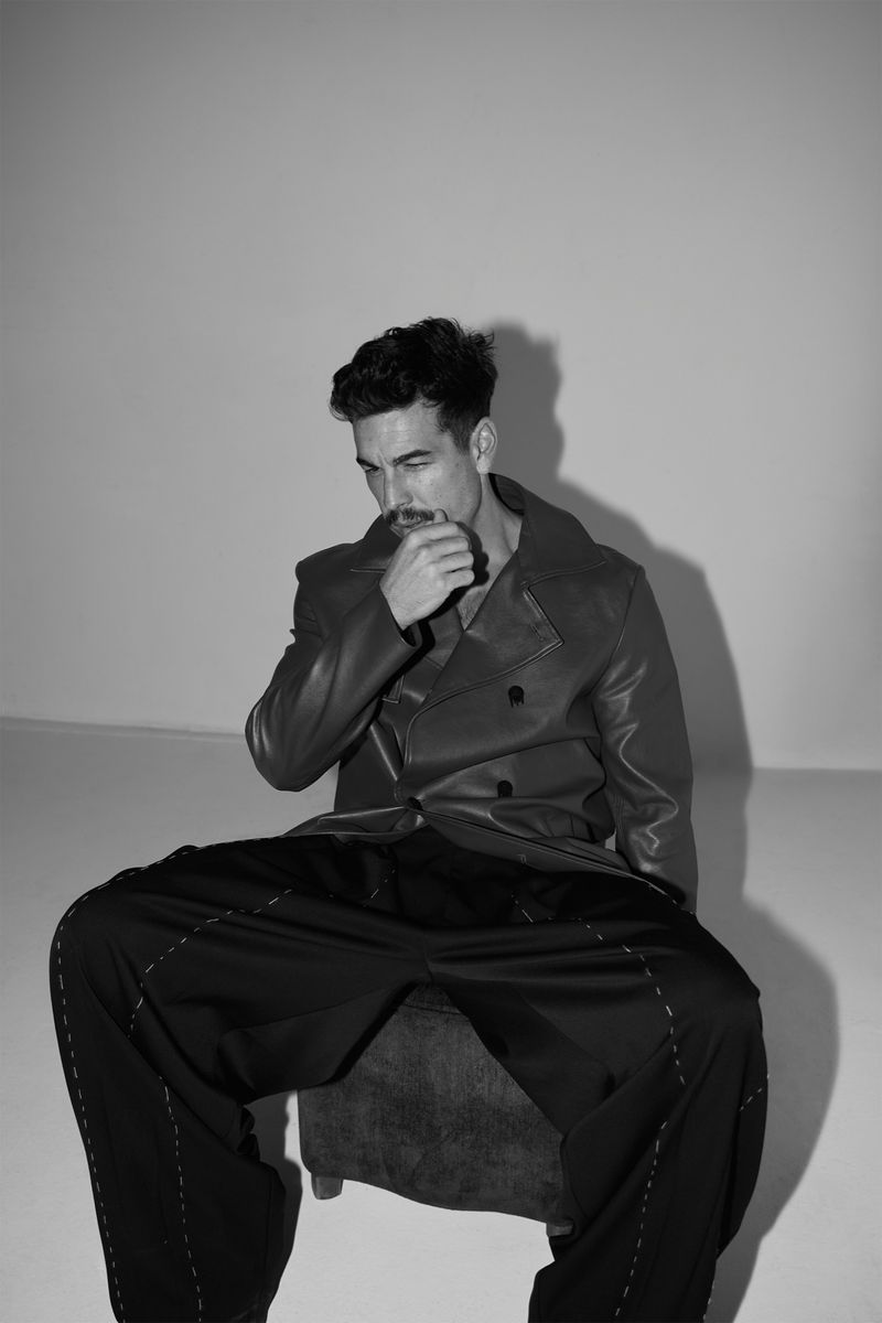 Monochrome portrait of a man in an overcoat sitting on a chair, hand to chin, looking down in a brightly lit photo studio setting.