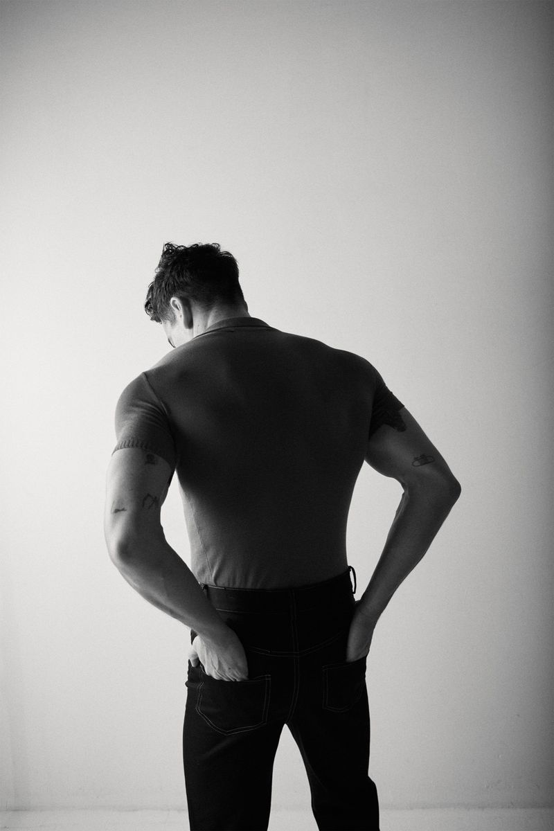 Black and white shot of a man facing away from the camera wearing a t-shirt and jeans, his hands in his pockets, against a white wall.