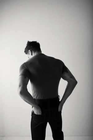 Black and white shot of a man facing away from the camera wearing a t-shirt and jeans, his hands in his pockets, against a white wall.