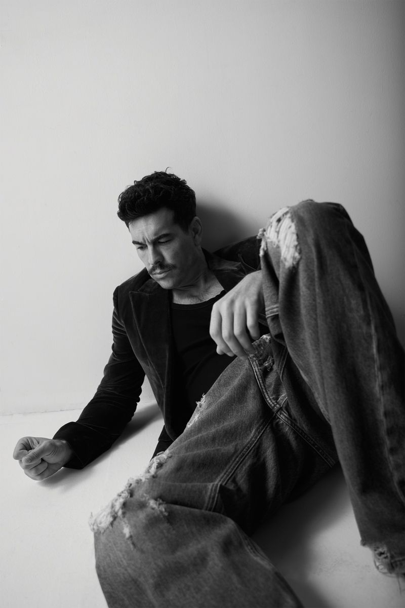 Monochrome portrait of an adult male posing indoors with ripped jeans, a black top, and a jacket against a light-colored wall.