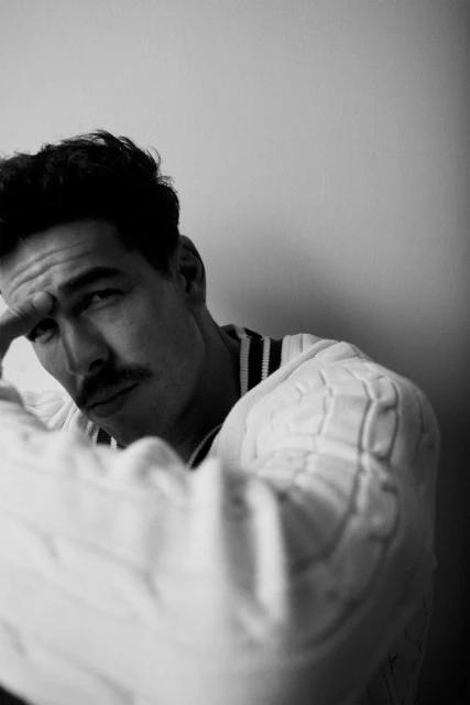 Monochrome portrait of an adult man with dark hair and a mustache, wearing a cable-knit sweater, posing indoors against a white wall.