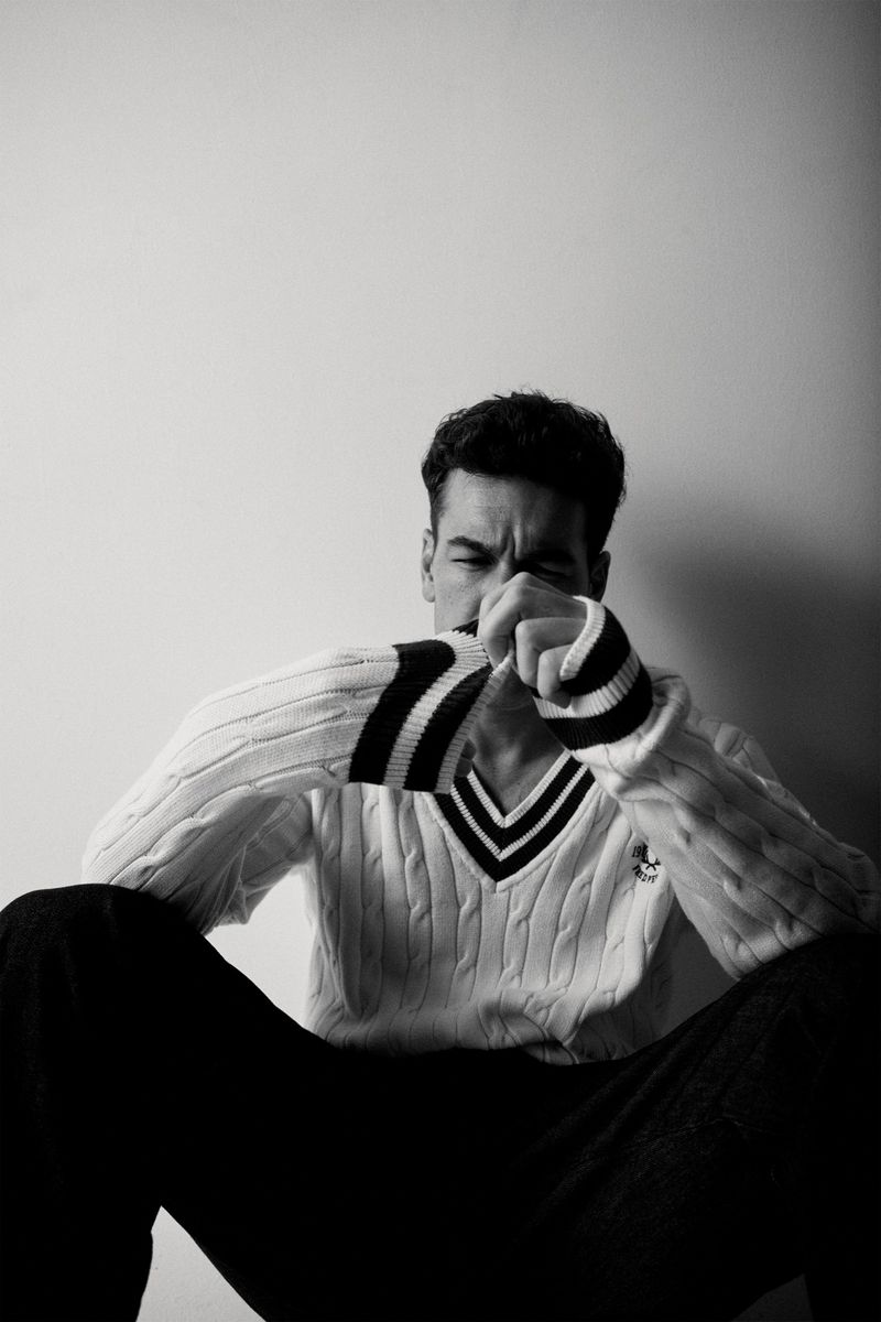 Monochrome portrait of a young adult man seated indoors wearing a sweater, posed in front of a plain wall, artistic and casual.