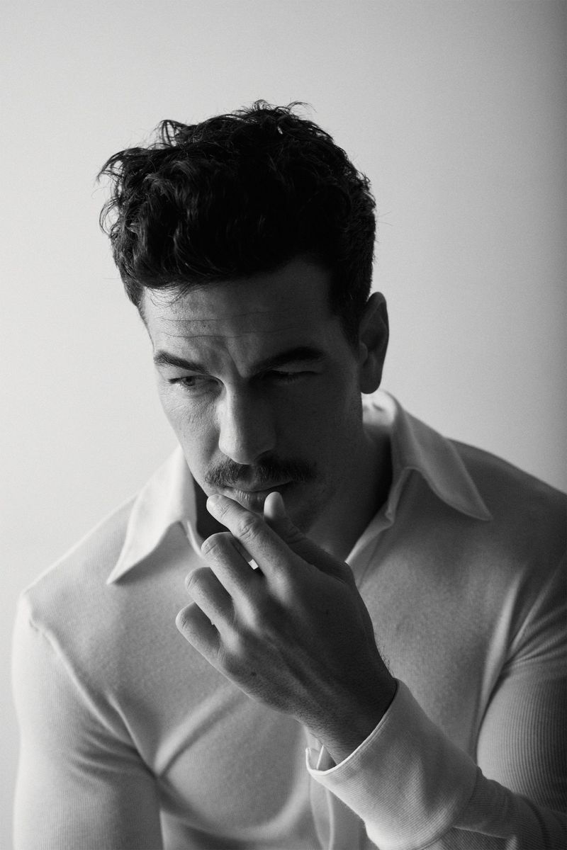 Black and white portrait of an adult male with dark hair and a mustache, wearing a collared shirt, touching his lip in an indoor setting.