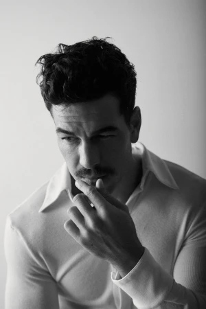 Black and white portrait of an adult male with dark hair and a mustache, wearing a collared shirt, touching his lip in an indoor setting.