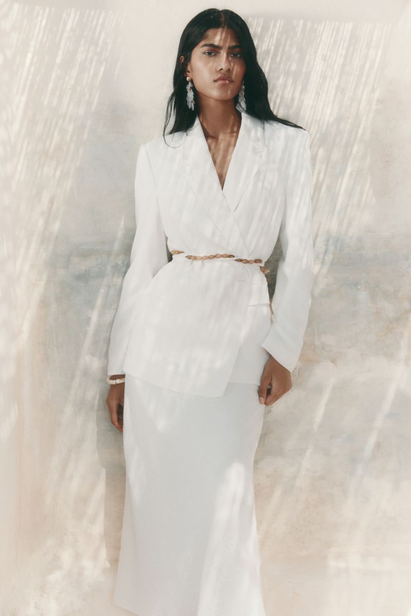A South Asian woman with long dark hair wears a white double-breasted blazer and matching maxi skirt, cinched with a thin golden braided waist belt.