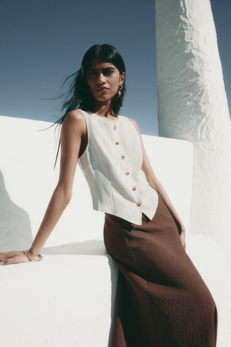 Model wearing a white buttoned vest top and brown skirt posing outdoors against stark white architectural structures.