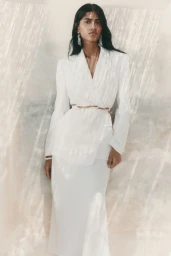 Model Ashley Radjarame wearing a white belted blazer and matching long skirt against a sun-dappled textured wall.