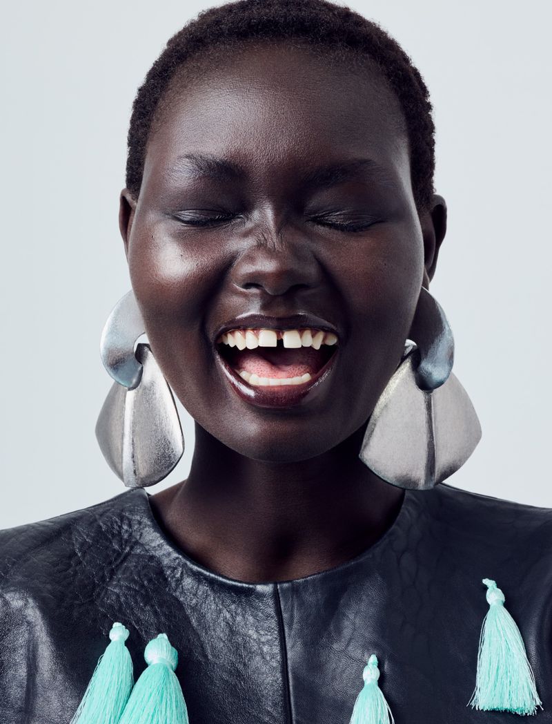 Close-up portrait of model Ateta Jok laughing with eyes closed, wearing large sculptural silver earrings and a black top with mint tassels.