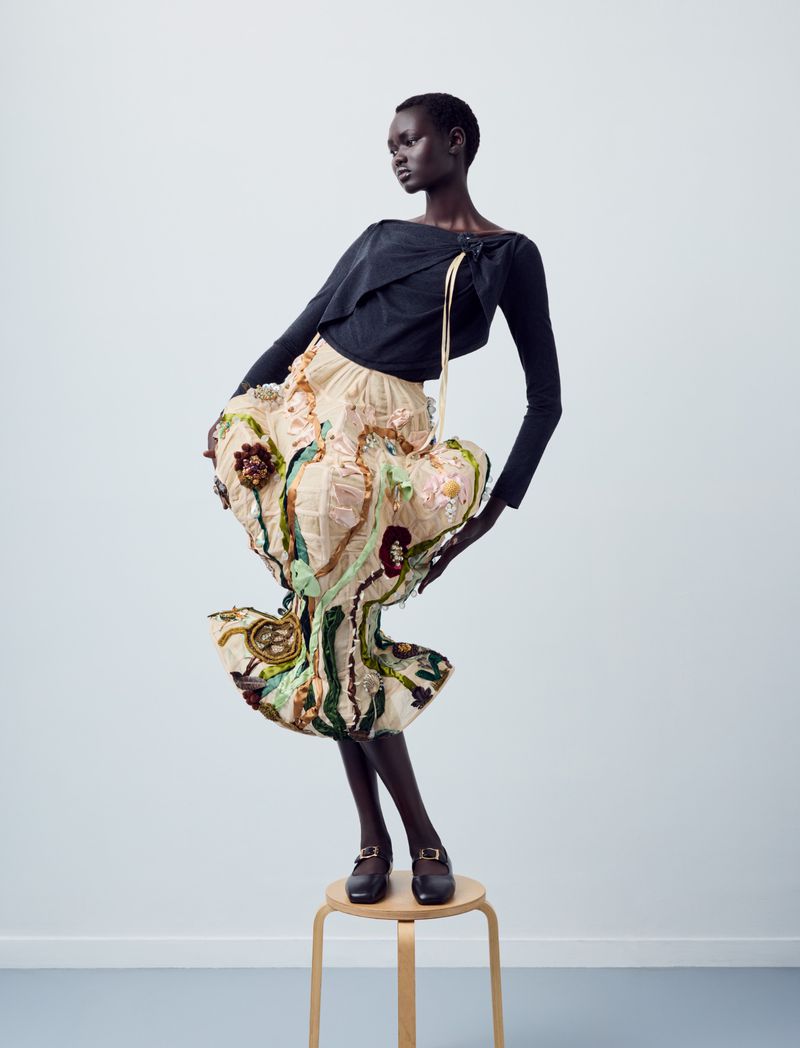 A model standing on a wooden stool wearing a black top and a beige skirt with intricate, colorful floral appliqué and structured draping.