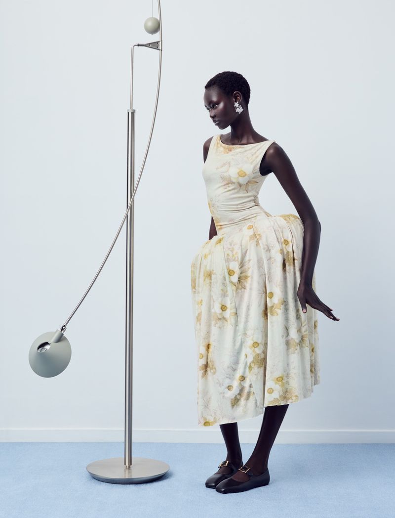 Ateta Jok posing in an ivory floral dress with a structured skirt beside a modern metal floor lamp in a minimalist studio setting.