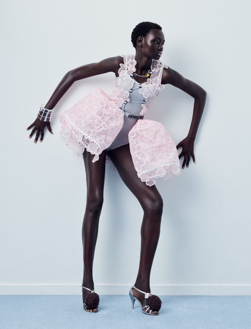 Model Ateta Jok in a structured pink lace mini dress and pom-pom heels posing against a pale studio wall.
