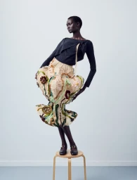 Model Ateta Jok poses atop a wooden stool wearing a sculptural embroidered skirt and black top against a white studio background.