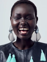 Close-up portrait of model Ateta Jok laughing with eyes closed, wearing large sculptural silver earrings and a black top with mint tassels.