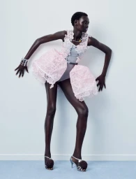 Model Ateta Jok in a structured pink lace mini dress and pom-pom heels posing against a pale studio wall.