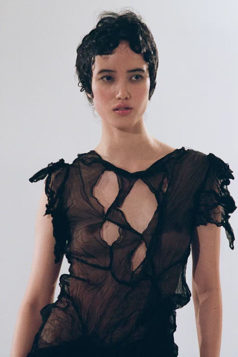 Close-up portrait of a woman with short dark curly hair wearing a sheer black distressed top with cut-out details against a plain background.