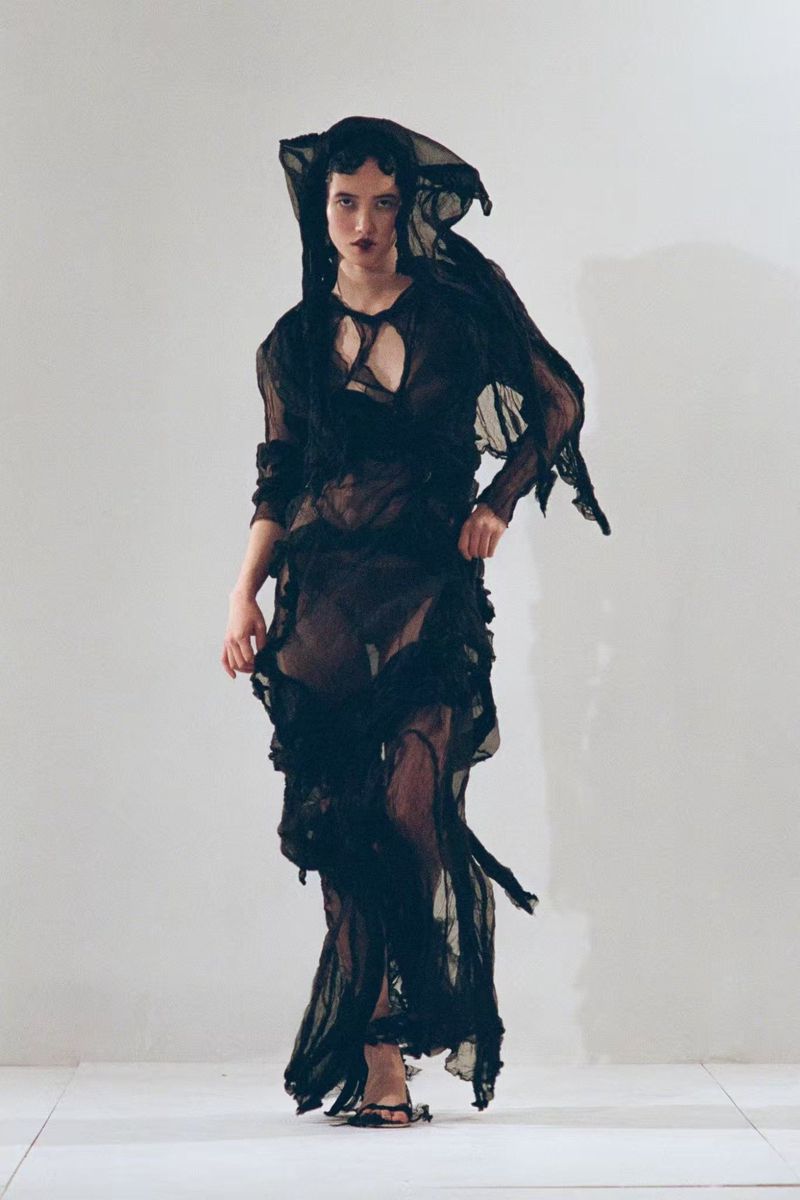 Model Yidan Huang wearing a sheer black deconstructed hooded gown against a light studio backdrop for Paolo Carzana.