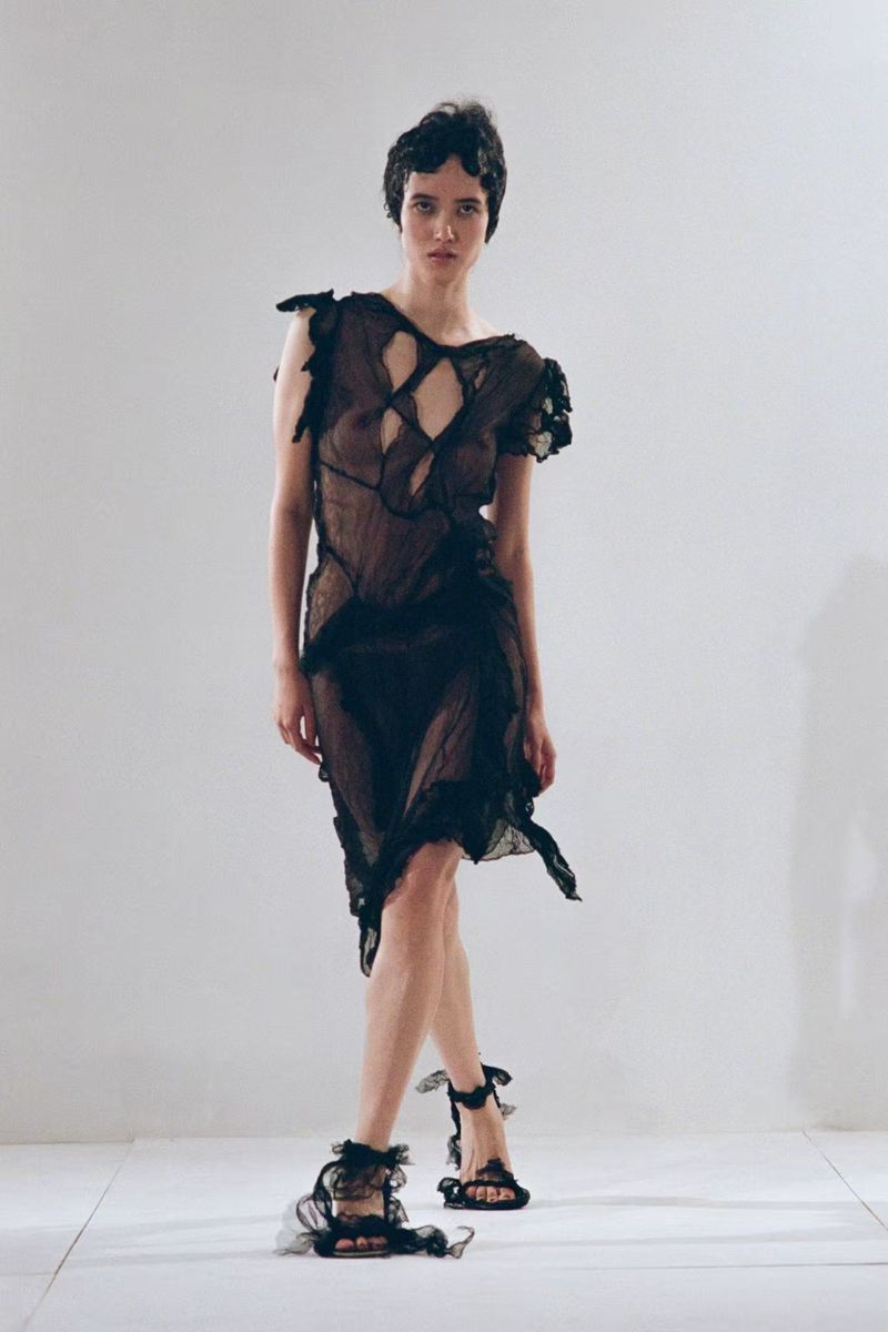 Model wearing a sheer black cutout midi dress and ruffled black sandals against a white studio backdrop.