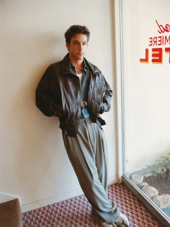 Role Model posing in an oversized brown leather jacket and pleated wide-leg trousers leaning against an indoor wall by a window.