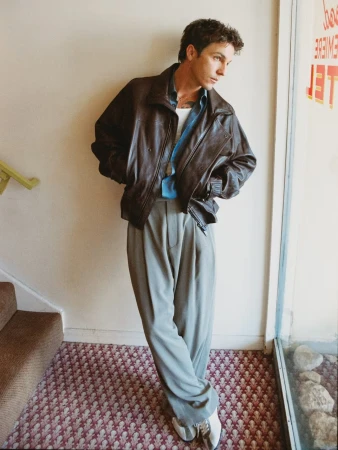 Musician Role Model leaning against an off-white wall wearing an oversized brown leather jacket and wide-leg grey trousers.