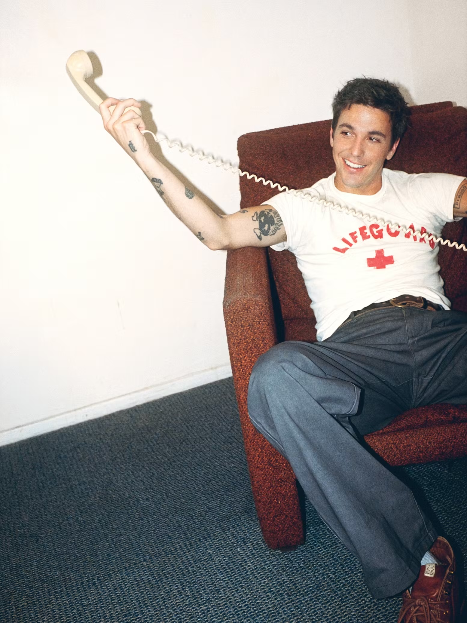 A smiling young man with arm tattoos relaxes in a brown armchair holding a cream telephone receiver against a plain white wall.