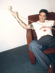 A smiling young man with arm tattoos relaxes in a brown armchair holding a cream telephone receiver against a plain white wall.