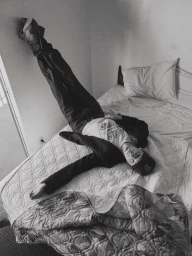 Black and white photo of a man lying on a bed with his legs propped straight up against the wall wearing a graphic tee and cap.